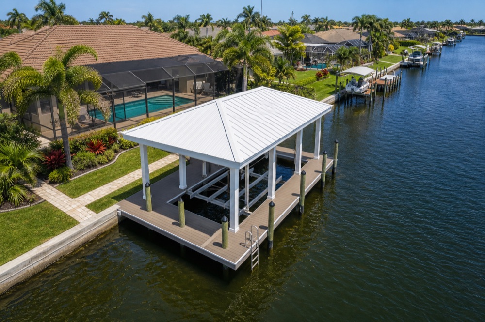 Aerial view of a white-roofed boathouse standing over an empty boat lift on a Southwest Florida canal, with a screened pool cage and landscaped lawn behind it and more waterfront homes down the water
