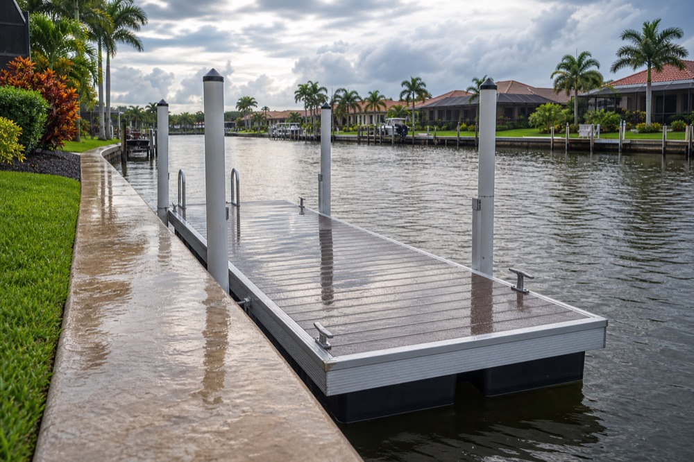 A floating dock moored alongside a concrete seawall under heavy grey overcast, tall guide piles standing at each corner and the decking still wet