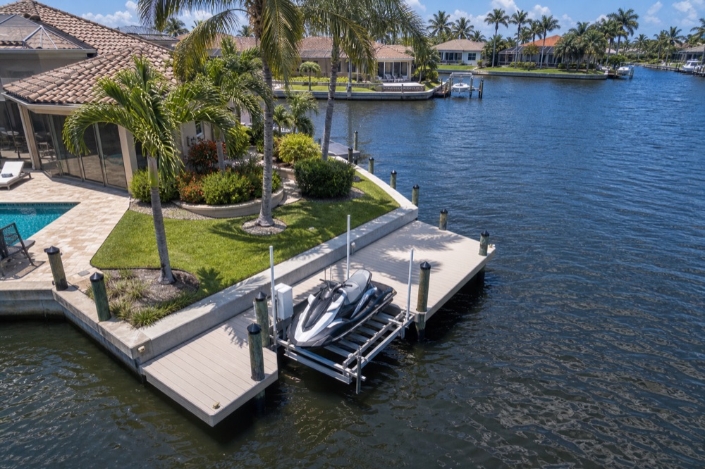 Aerial view of a waterfront home with a pool and paver deck on a point lot, a personal watercraft raised on an aluminum jet ski lift at the composite dock, and a wide canal of homes on the far bank