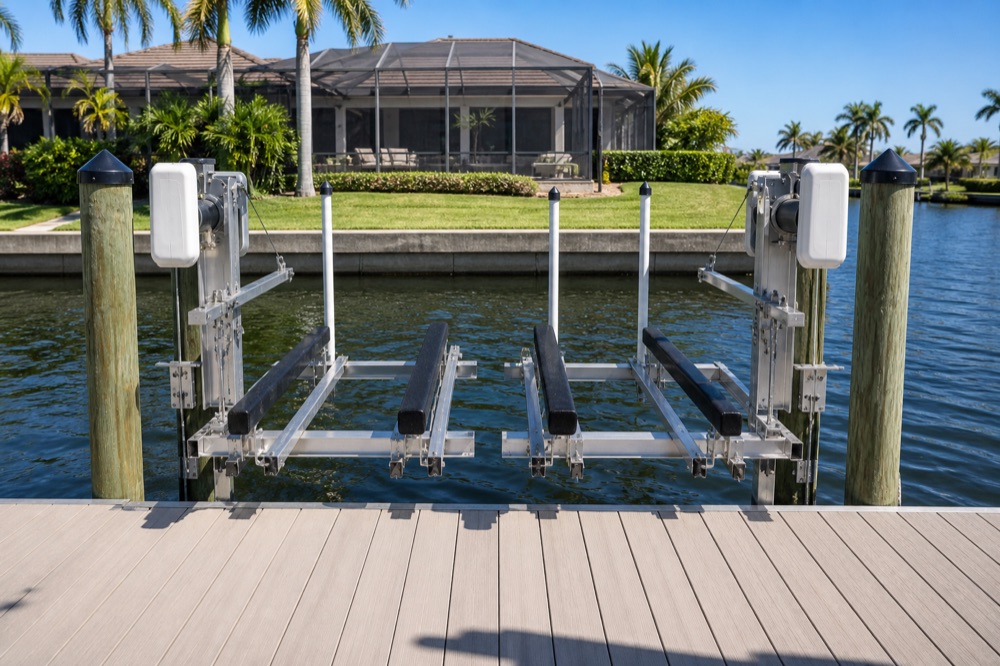 An empty aluminum boat lift with black bunk beams suspended over the water between capped pilings, with a screened lanai and lawn behind