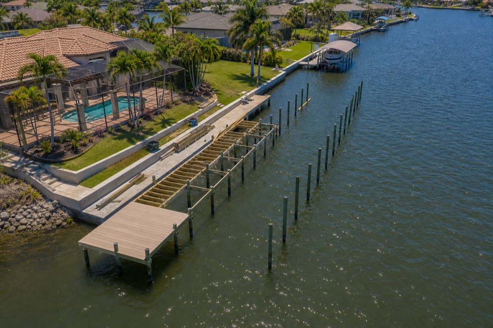 Aerial view of a waterfront home and pool beside a long seawall, with a dock carrying boat lift framing and a row of newly driven pilings following a curving canal