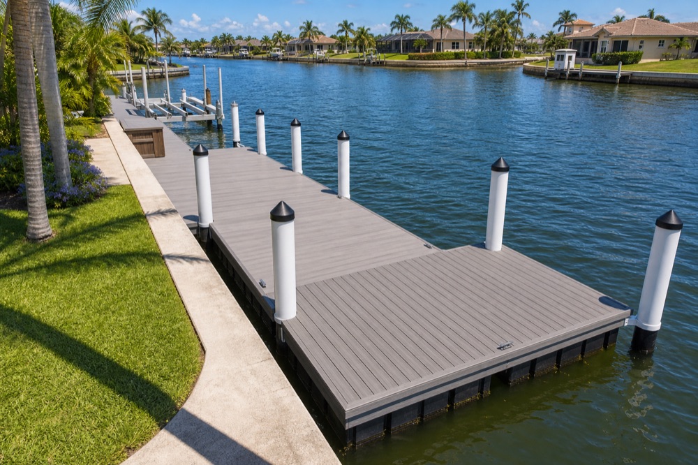 Elevated view of a gray composite dock walkway running along a concrete seawall out to a wide platform, with white-wrapped pilings under black conical caps, lift beams further up the walkway, and a wide canal with homes and palms on the far bank